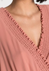 Růžová blůzka s výstřihem do V, se strukturovanými smykovými detaily, volánkovými akcenty kolem výstřihu a měkkou, lehkou tkaninou.