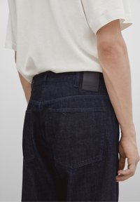 Tummansiniset denimhelsit, joissa on hieman kapeneva lahje, hienovarainen tekstuuri ja kaksi takataskua. Yhdistetty vaaleanväriseen t-paitaan.