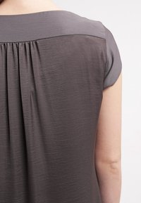 Blusa gris de manga corta, con una textura suave y detalles fruncidos en la parte posterior. El tejido parece liviano y cae con suavidad.