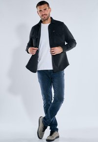 Mann in einer schwarzen Jacke über einem weißen Hemd, blauen Jeans und hellgrauen Turnschuhen, der mit gekreuzten Beinen vor einem einfarbigen Hintergrund steht.