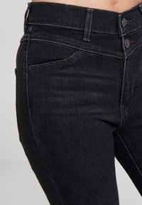 Pantalones vaqueros de mezclilla negros con corte ajustado, con un cierre de dos botones, bolsillos frontales y un sutil detalle de costura a lo largo de las costuras.