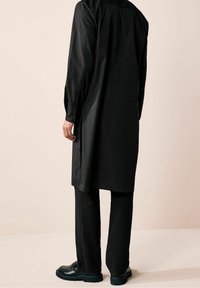 Chemise longue noire à coupe droite, détails de boutons sur les manches, associée à un pantalon noir et des chaussures noires. Texture lisse, design minimaliste.