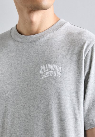 Prim-plan cu o persoană care poartă un tricou gri cu text alb "Billionaire Boys Club" și logo-ul unui astronaut pe piept.