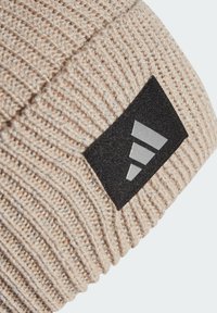 Berretto a maglia beige con una texture a costine, caratterizzato da un'etichetta nera rettangolare con tre strisce bianche su un lato.