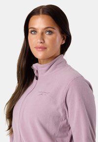 Purppura fleece-takki, jossa on korkea kaulus ja vetoketjukiinnitys, jossa on hienovaraisesti kohokuvioitu logo rinnassa ja pehmeä, rakennepintainen viimeistely.