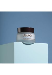 Ahava Active Moisture Gel Cream i en frostad glasburk med ett mörkbrunt lock, placerad på en ljusblå geometrisk yta, mot en blå bakgrund.