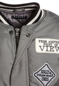 Šedá bomber bunda s černým pruhovaným límcem. Obsahuje zipy, vyšívané nášivky a hladký, texturovaný povrch.