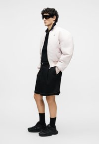 Homme portant une veste oversize rose clair, un t-shirt noir, un short noir, des chaussettes noires, des baskets noires et de grandes lunettes de soleil noires, debout les mains dans les poches.