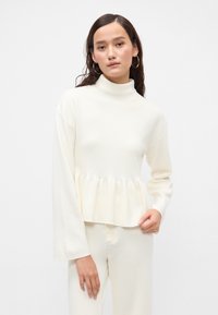 Top bianco a maniche lunghe con colletto alto e design a peplo. Texture morbida, vestibilità rilassata e polsini a costine. Abbinato a pantaloni coordinati.