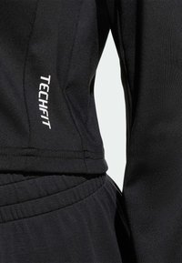 Čierny atletický top s hladkou, elastickou textúrou a priliehavým strihom. Biela potlač loga "TECHFIT" na bočnom šve.