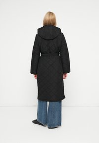 Juoda pūsta ilga striukė su kapišonu ir diržu, su deimanto raštu. Laisvas kirpimas, rodoma iš nugaros, derinama su plačiomis džinsų kelnėmis.