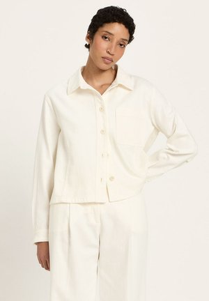 Young person with short curly hair wearing an off-white buttoned shirt and matching wide-leg pants, standing with one hand on hip.