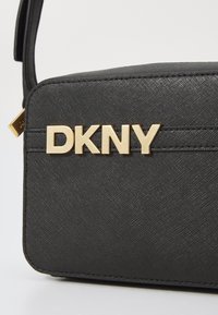 Melnā sintētiskā rokassoma ar struktūras virsmu, taisnstūrveida formu un zelta "DKNY" logo uz priekšpuses. Iekļauta regulējama siksna.