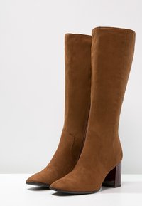 Bottes hauteur genou en daim marron avec un bout pointu et un talon épais, dotées d'une fermeture éclair latérale et d'une texture lisse.