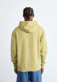 Persona con capelli intrecciati che indossa una felpa con cappuccio verde chiaro e jeans blu, vista di spalle su uno sfondo bianco semplice.