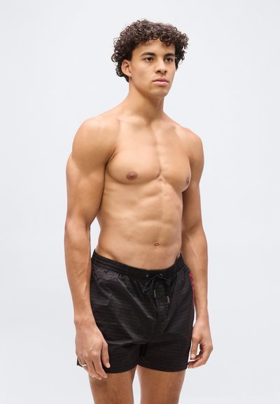 Νεαρός άνδρας με σγουρά μαλλιά που φοράει μαύρο μαγιό, όρθιος μπροστά σε ένα απλό ανοιχτό γκρι φόντο.