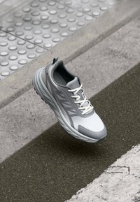Lightweight athletic shoe in gray and white, with a mesh upper, textured sole, and contrasting rubber accents. Features flat laces and a rear pull tab.