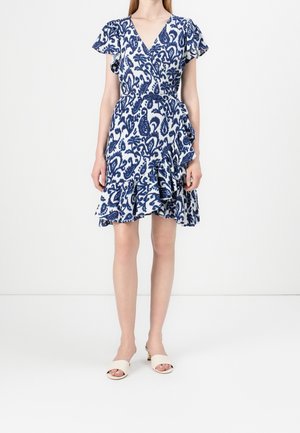 Femme portant une robe portefeuille à motifs bleu et blanc avec un ourlet à volants et des manches courtes, assortie à des sandales blanches à talons et à bout ouvert.
