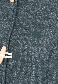 Detailní záběr na tmavozelenou tkaninu s tečkami, která má dřevěný knoflík a bílou šňůrku na oblečení.