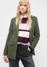 Olivovo-zelený blazer so štruktúrovaným dizajnom, ktorý má jedno vrecko, preložený cez pruhovaný sveter v ružovej, bielej a bordovej farbe, s čiernymi džínsami.