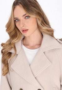 Beige dobbeltradet frakke med brede revers og messingknapper. Blødt, tekstureret stof; båret over en hvid top med klassisk kravedesign.