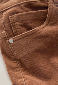 Tissu en velours côtelé marron avec des côtes verticales ; présente un design de poche classique avec un détail de bouton en métal argenté. Surface texturée.