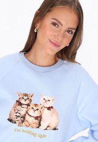 Világoskék pulóver, amelyen három masnis kiscica grafikája és arany színű "I'm holding tight" felirat található. Puha tapintású és laza szabású.