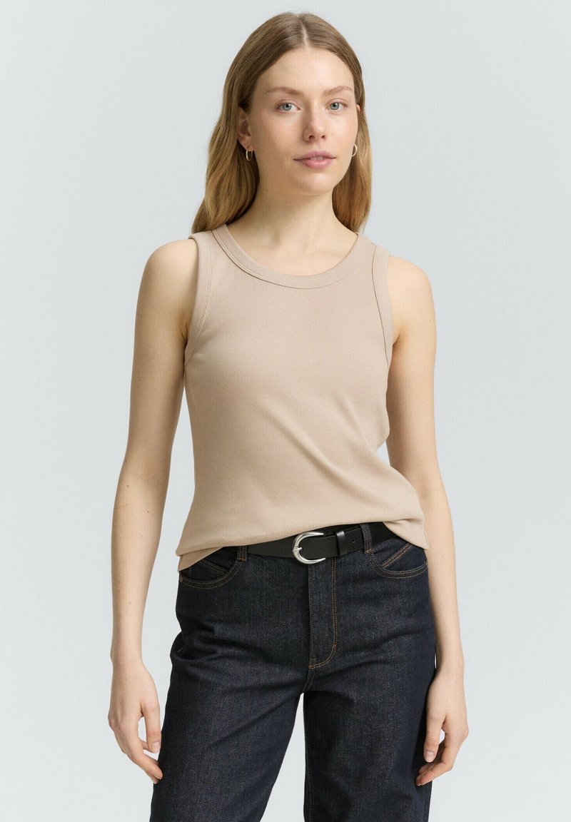 Top sans manches beige en tissu côtelé, avec un col rond et une coupe classique, associé à un jean en denim foncé et une ceinture noire.