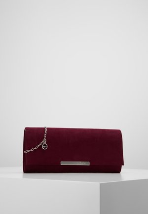 Bolso clutch rectangular de ante rojo oscuro con cadena plateada y pequeño charm redondo, descansando sobre una superficie blanca frente a un fondo liso.
