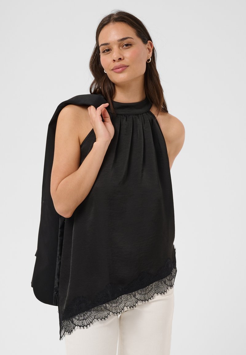 Femme portant un haut noir sans manches à col montant orné de dentelle, un pantalon blanc et une veste noire posée sur son épaule, debout devant un fond uni.
