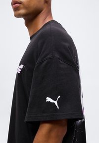 Άνδρας με μαύρο μπλουζάκι Puma με λευκό λογότυπο Puma στο μανίκι, παρουσιασμένος από το πλάι σε απλό φόντο.