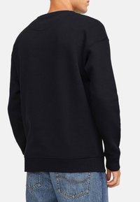 Felpa nera con collo rotondo, polsini e orlo a coste, realizzata in morbido tessuto a trama semplice, indossata con jeans azzurri chiari.