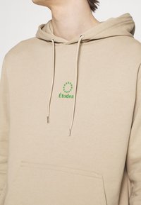 Beżowa bluza z kapturem wykonana z miękkiego materiału, z kieszenią z przodu, regulowanymi sznurkami zakończonymi metalowymi końcówkami oraz zielonym haftowanym logo.