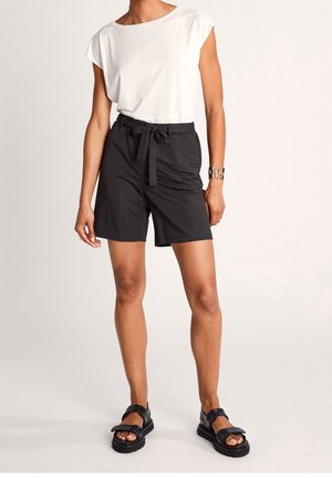 Mujer que lleva una blusa sin mangas blanca, pantalones cortos negros con cinturón, sandalias negras y una pulsera de metal plateada, de pie sobre un fondo claro.