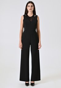 Jumpsuit senza maniche nero con inserti in pizzo, taglio sartoriale, gamba ampia e pieghe sottili. Indossato con tacchi neri lucidi.