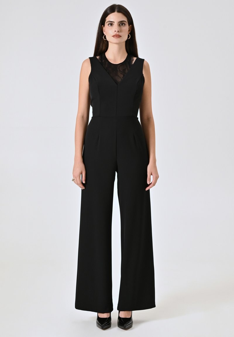Jumpsuit senza maniche nero con inserti in pizzo, taglio sartoriale, gamba ampia e pieghe sottili. Indossato con tacchi neri lucidi.