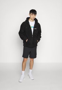 Svart fleece zip-up hettegenser med hette, to frontlommer, brukt over en hvit t-skjorte, sammen med mørke shorts og hvite joggesko.