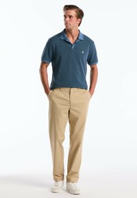 Polo shirt bleu à texture avec des manches courtes, col contrasté, associé à un pantalon beige et des baskets blanches, se tenant devant un fond blanc.