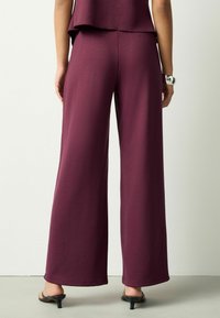 Bordové nohavice s širokými nohavicami vyrobené z mäkkej tkaniny s hladkým povrchom a elastickým pásom. Nosia sa s čiernymi sandálmi na podpätku.