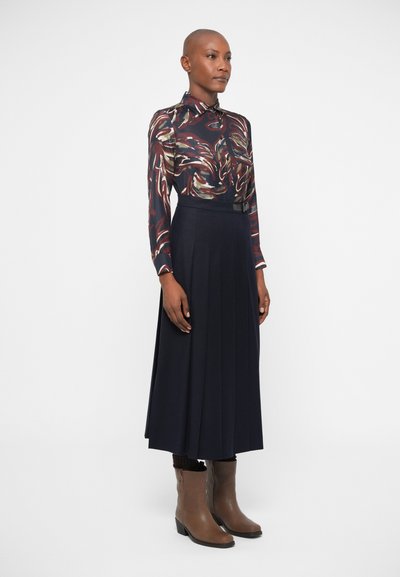 Mönstrad blus med lång ärm och svängande mönster i mörka nyanser, i kombination med en plisserad marinblå kjol och bruna ankla stövlar med klack.