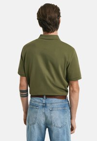 Olivgrön polo med kort ärm, tillverkad av slät tyg, med en klassisk krage. Agerad med ljusblå denimjeans.
