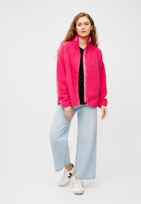 Giacca rosa pelosa con colletto alto e zip frontale, abbinata a un top nero e jeans a gamba larga blu chiaro. Sneakers bianche con accentuazioni nere.