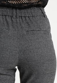 Černé a šedé kostkované kalhoty s elastickým pasem a bočními kapsami. Tkanina má strukturovanou vazbu. Detailní pohled ukazující designové prvky.