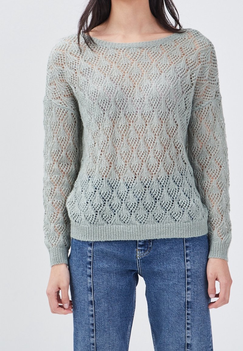 Femme portant un pull en dentelle tricotée vert clair à manches longues, associé à un jean en denim bleu, debout devant un fond uni.