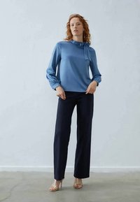 Lyseblå bluse med en knyttet detaljer ved halsen, lange ærmer og en glat tekstur, parret med marineblå tapered bukser og guld sandaler.