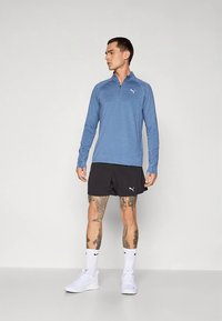 Gaiši zila garām piedurknēm krekls ar pusjostiņu un logo krūtīs, kopā ar melnām šortiem. Komplektā baltas sporta kurpes un zeķes.