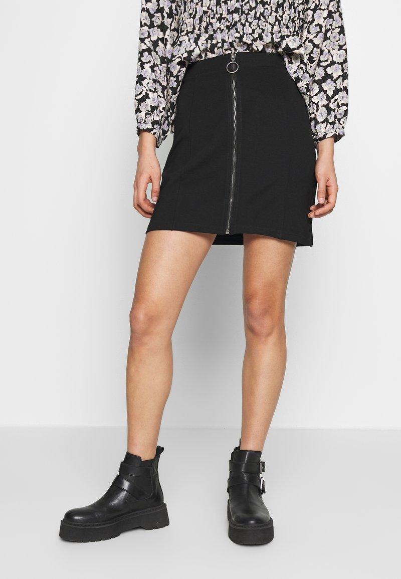 Femme portant une mini-jupe noire avec fermeture éclair à l'avant, un top à manches longues fleuri et des bottines noires, debout sur fond blanc.