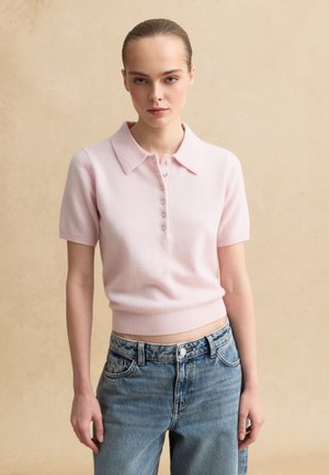 Donna in piedi con postura eretta che indossa una polo rosa pallido a maniche corte con bottoni e jeans a vita alta di colore azzurro chiaro.