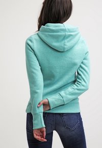 Felpa verde menta con tasca a marsupio, cappuccio regolabile con cordino e polsini a costine, abbinata a jeans in denim scuro.