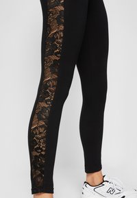 Leggings noirs avec un panneau latéral en dentelle noire élaborée, présentant des motifs floraux. Le tissu est extensible et moulant. Baskets blanches visibles.